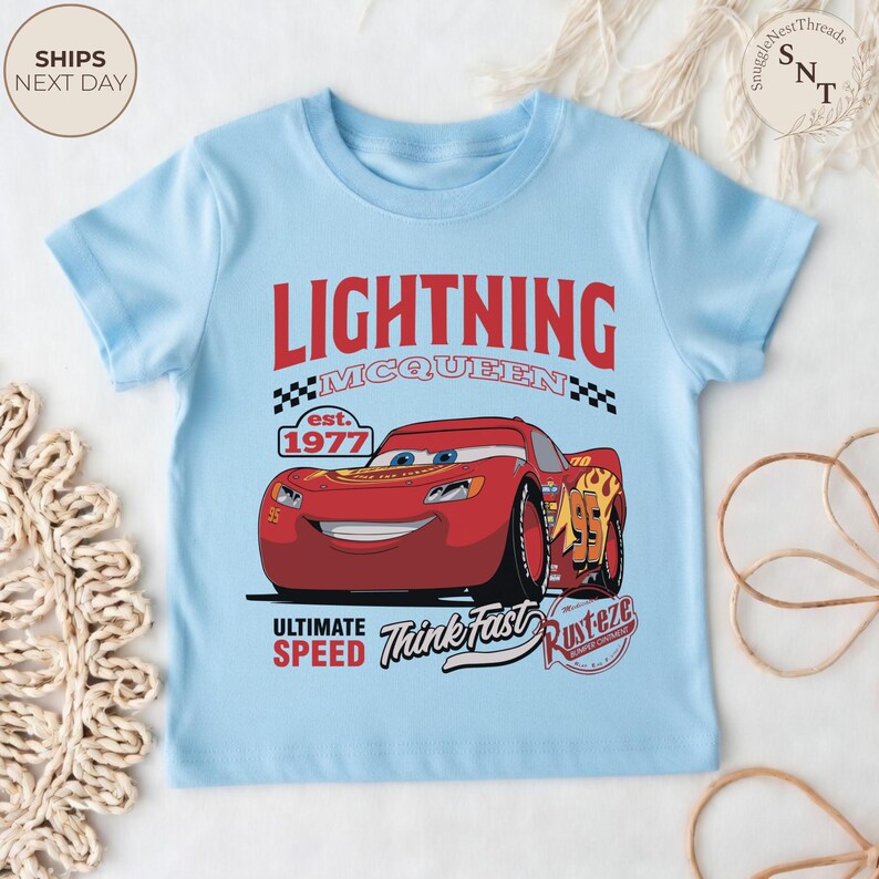 Op de afbeelding: Lichtblauw T-shirt met een afbeelding van Lightning McQueen, een rode raceauto, uit de film Cars. Het ontwerp bevat de tekst "LIGHTNING MCQUEEN" en "ULTIMATE SPEED". Het shirt heeft ook de tekst "Think Fast".