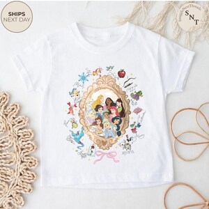 May include: White t-shirt with a gold-framed portrait of Disney princesses, surrounded by character illustrations and names. A pink bow is at the bottom. The shirt has short sleeves and a crew neck.