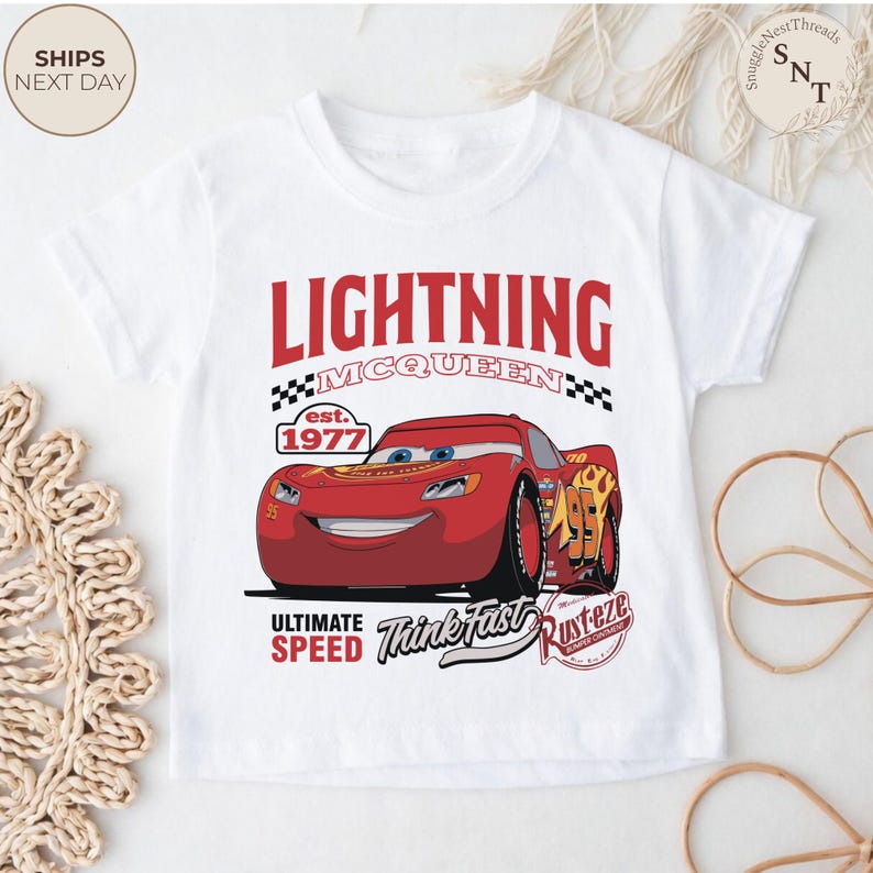 Op de afbeelding: Witte t-shirt met een Lightning McQueen-ontwerp. De rode raceauto-afbeelding wordt vergezeld door de tekst "LIGHTNING MCQUEEN", "est. 1977", "ULTIMATE SPEED" en "Think Fast". Een kledingstuk voor kinderen.