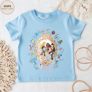 May include: A pale blue t-shirt with a gold-framed portrait of animated princesses. The design includes character illustrations and signatures. The shirt has short sleeves and a round neck.