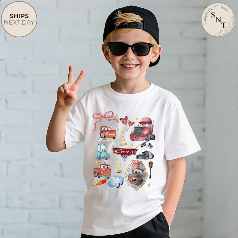 May include: White t-shirt featuring colorful illustrations of characters from the movie "Cars." The design includes images of race cars, trucks, and the word "Cars" in a red banner. The shirt is worn by a person wearing sunglasses and a black cap.