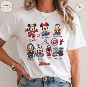 May include: White t-shirt featuring a colorful graphic of Disney characters as Avengers. The design includes Mickey Mouse, Minnie Mouse, Donald Duck, Daisy Duck, Goofy, Pluto, Chip & Dale, and the Avengers logo. The shirt also has the text "SnuggleNest Threads".