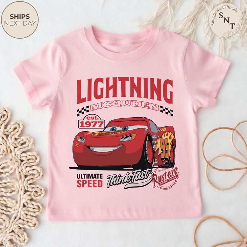 Op de afbeelding: Een lichtroze t-shirt met een afbeelding van Lightning McQueen uit de film Cars. Het ontwerp bevat de tekst "LIGHTNING MCQUEEN" in het rood, het nummer "95" en de zinnen "ULTIMATE SPEED" en "Think Fast".