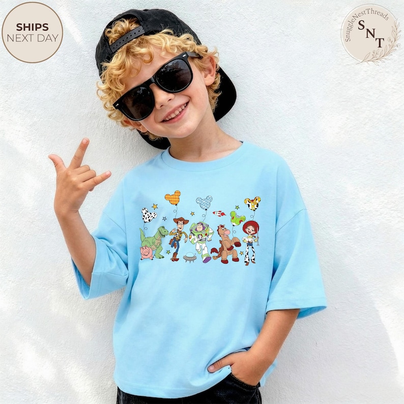 May include: Light blue t-shirt featuring a colorful graphic of Toy Story characters. The design includes Woody, Buzz Lightyear, and other characters from the movie. The shirt has short sleeves and a relaxed fit, perfect for casual wear.