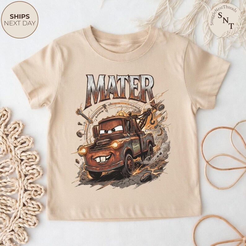Puede incluir: Camiseta beige de manga corta con un gr&aacute;fico de Mate de la pel&iacute;cula Cars. La gr&uacute;a marr&oacute;n tiene una cara sonriente, rodeada de herramientas y la palabra "MATER". La camiseta tiene cuello redondo.