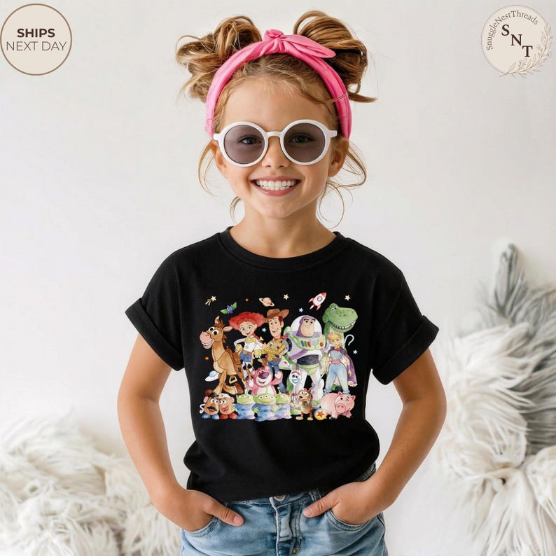 May include: A black t-shirt featuring a colorful graphic of characters from the animated movie "Toy Story". The design includes Woody, Buzz Lightyear, and other beloved characters. The shirt is worn by a young person.