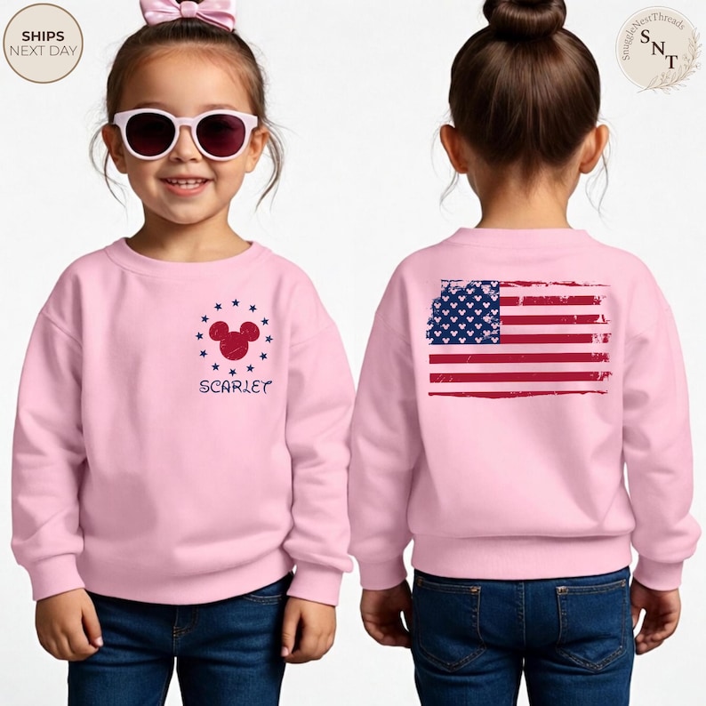 May include: Pink sweatshirt featuring a Mickey Mouse design with stars and the name "Scarlet" in navy blue. The back of the sweatshirt displays a distressed American flag graphic. The child is wearing blue jeans.
