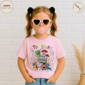 May include: A light pink t-shirt featuring the words "TOY STORY" above a colorful graphic of characters from the animated movie. The shirt is worn by a child wearing heart-shaped sunglasses and has black hair ties.