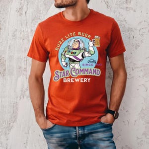 Puede incluir: Camiseta de color rojo anaranjado con un gr&aacute;fico de dibujos animados de Buzz Lightyear sosteniendo una jarra de cerveza. El dise&ntilde;o incluye el texto "Buzz Lite Beer" y "Star Command Brewery" en un estilo vintage. La camiseta es de cuello redondo y manga corta.