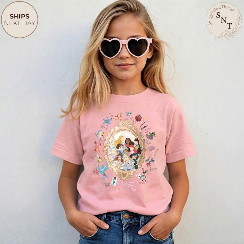 May include: Pale pink t-shirt with a framed illustration of Disney princesses. The shirt has a crew neck and short sleeves. The image includes the text "Ships Next Day" in a circle on the upper left.