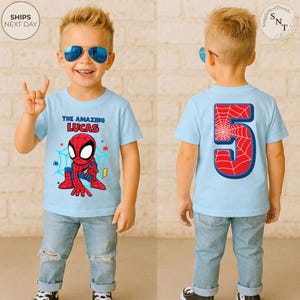 Puede incluir: Camiseta azul claro con tem&aacute;tica de Spider-Man con el texto "THE AMAZING LUCAS" y un gr&aacute;fico de Spider-Man. La parte posterior de la camiseta presenta el n&uacute;mero 5 con un dise&ntilde;o de telara&ntilde;a. El ni&ntilde;o lleva vaqueros azules.
