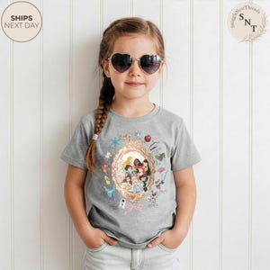 May include: A gray t-shirt featuring a gold-framed illustration of Disney princesses. The shirt has a crew neck and short sleeves. The text "SHIPS NEXT DAY" is in a circle on the top left.