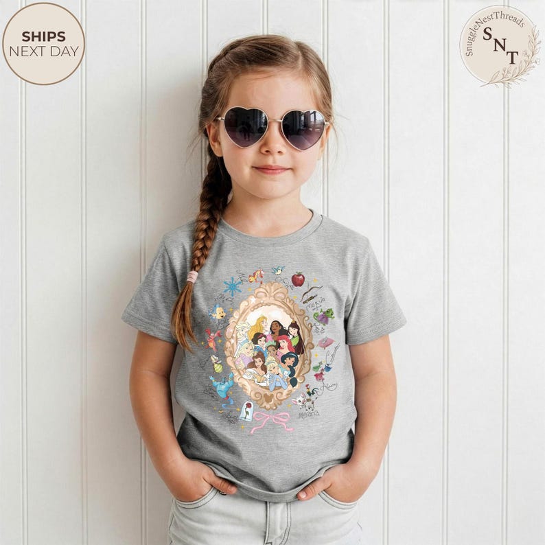 May include: A gray t-shirt featuring a gold-framed illustration of Disney princesses. The shirt has a crew neck and short sleeves. The text "SHIPS NEXT DAY" is in a circle on the top left.