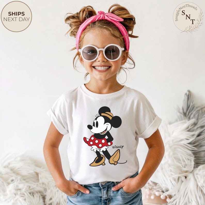May include: A white t-shirt featuring a Minnie Mouse graphic with a red and white polka dot skirt and the Disney logo. The child is wearing a pink headband and sunglasses. The image includes the text "SHIPS NEXT DAY".
