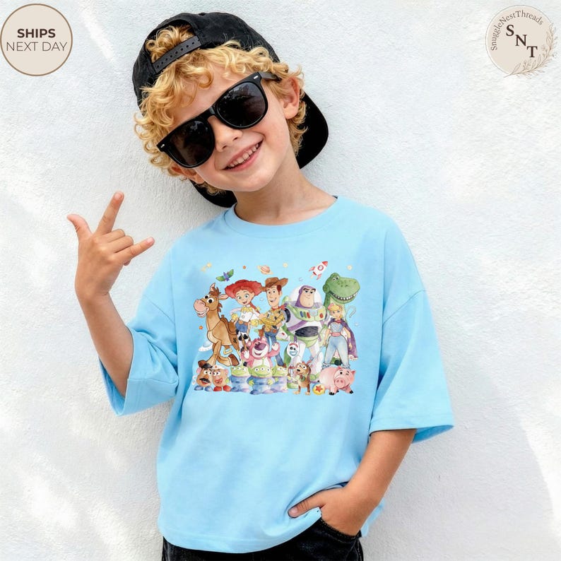 May include: Light blue t-shirt featuring a colorful graphic of characters from the animated movie Toy Story. The shirt has short sleeves and a relaxed fit. The child in the image is wearing a black cap and sunglasses.