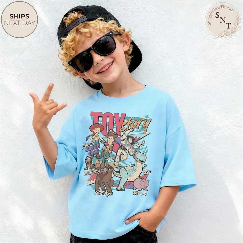 Puede incluir: Una camiseta azul claro con un estampado gr&aacute;fico colorido de los personajes de Toy Story, incluyendo a Woody, Buzz Lightyear, Jessie, Perdig&oacute;n, Slinky, Rex y Hamm, con el texto Toy Story y los nombres de los personajes en fuente retro.