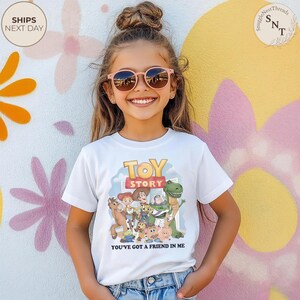 K&ouml;nnte beinhalten: Ein wei&szlig;es T-Shirt mit den Toy Story-Charakteren und dem Text "TOY STORY" und "YOU'VE GOT A FRIEND IN ME". Das T-Shirt wird von einer jungen Person mit rosa Sonnenbrille getragen.