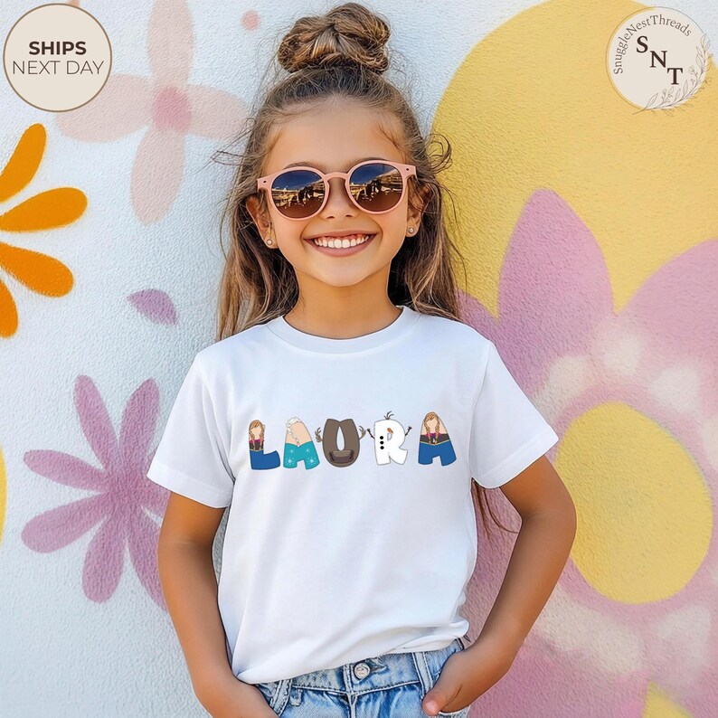 K&ouml;nnte beinhalten: Ein wei&szlig;es T-Shirt mit dem Namen "LAURA" in bunten Buchstaben aufgedruckt. Jeder Buchstabe ist mit einem anderen Charakterthema gestaltet. Das M&auml;dchen im Bild tr&auml;gt eine rosa Sonnenbrille und l&auml;chelt.