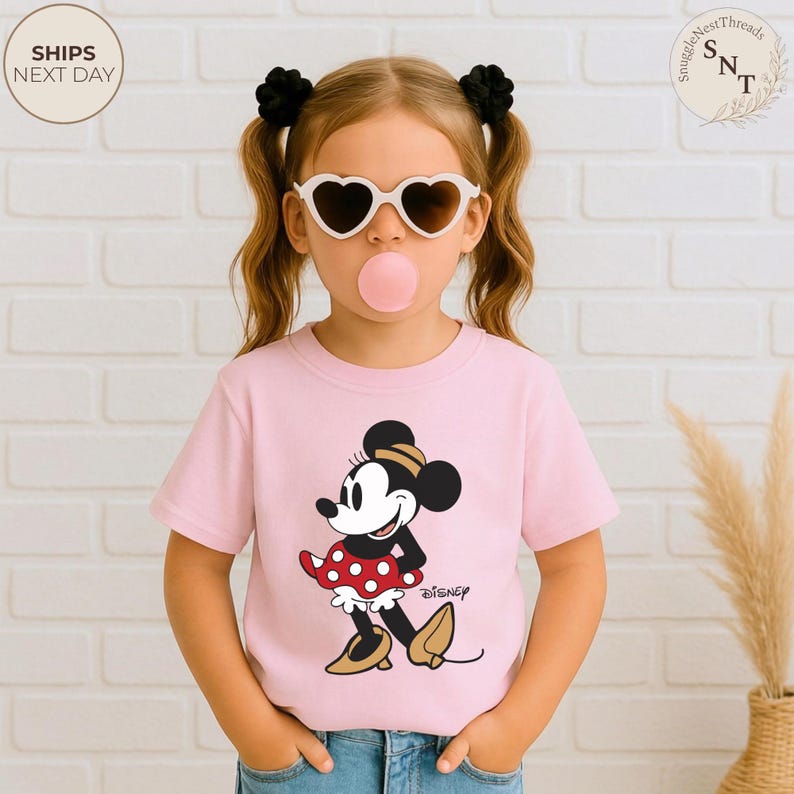 May include: A light pink t-shirt featuring a Minnie Mouse graphic. The cartoon character is wearing a red polka dot dress, black shoes, and a black hat. The word "Disney" is printed below the graphic. The child is wearing heart-shaped sunglasses and has a bubble gum bubble.