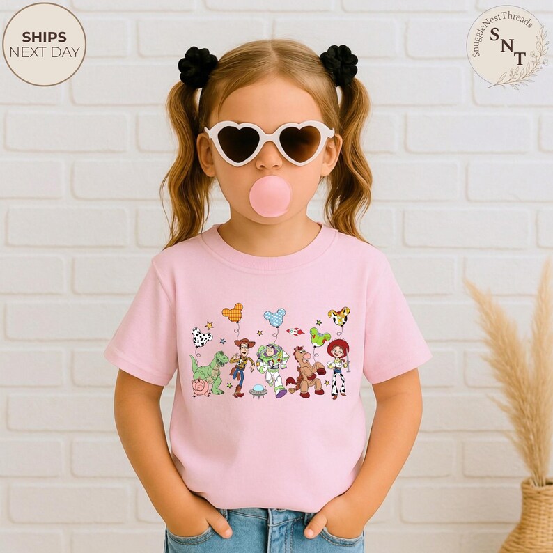 May include: A light pink t-shirt featuring a colorful graphic of cartoon characters and balloons. The design includes a dinosaur, a cowboy, a space ranger, a horse, and a cowgirl. The shirt is worn by a young person wearing heart-shaped sunglasses.