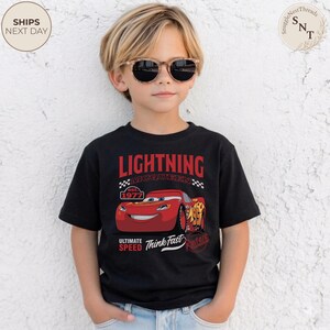 Op de afbeelding: Zwart Lightning McQueen T-shirt met rode tekst en een afbeelding van het personage. Het shirt heeft ook de tekst "Ultimate Speed Think Fast" en "est. 1977". Gedragen door een kind met een zonnebril.