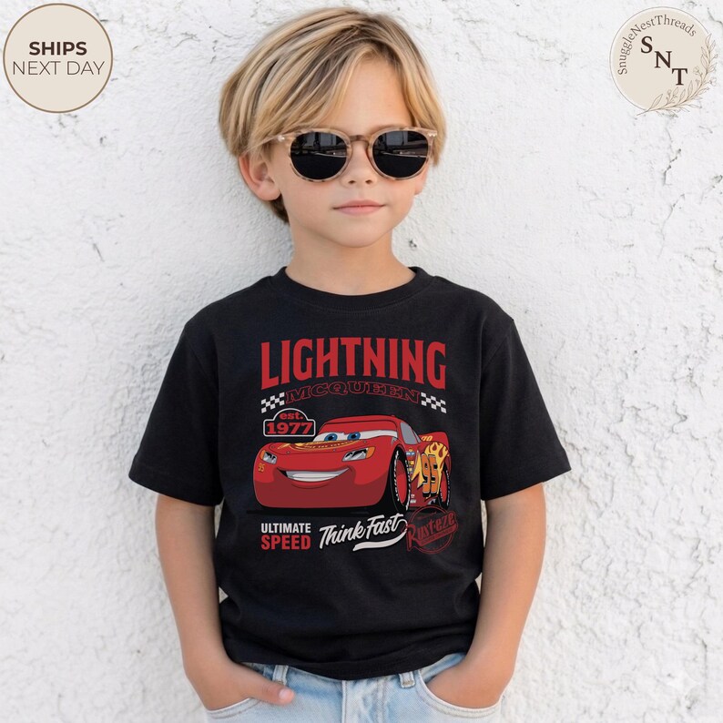 Op de afbeelding: Zwart Lightning McQueen T-shirt met rode tekst en een afbeelding van het personage. Het shirt heeft ook de tekst "Ultimate Speed Think Fast" en "est. 1977". Gedragen door een kind met een zonnebril.