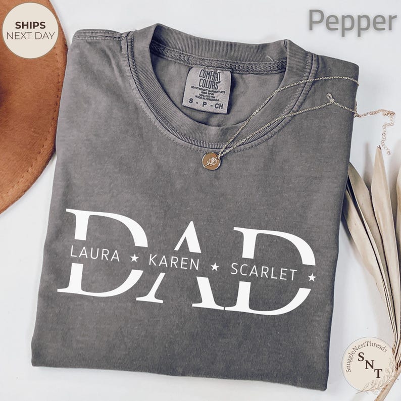 Puede incluir: Una camiseta Comfort Colors color pimienta con la palabra "DAD" en letras blancas grandes. Debajo, los nombres "LAURA", "KAREN" y "SCARLET". Una cadena de plata con un colgante est&aacute; sobre la camiseta.