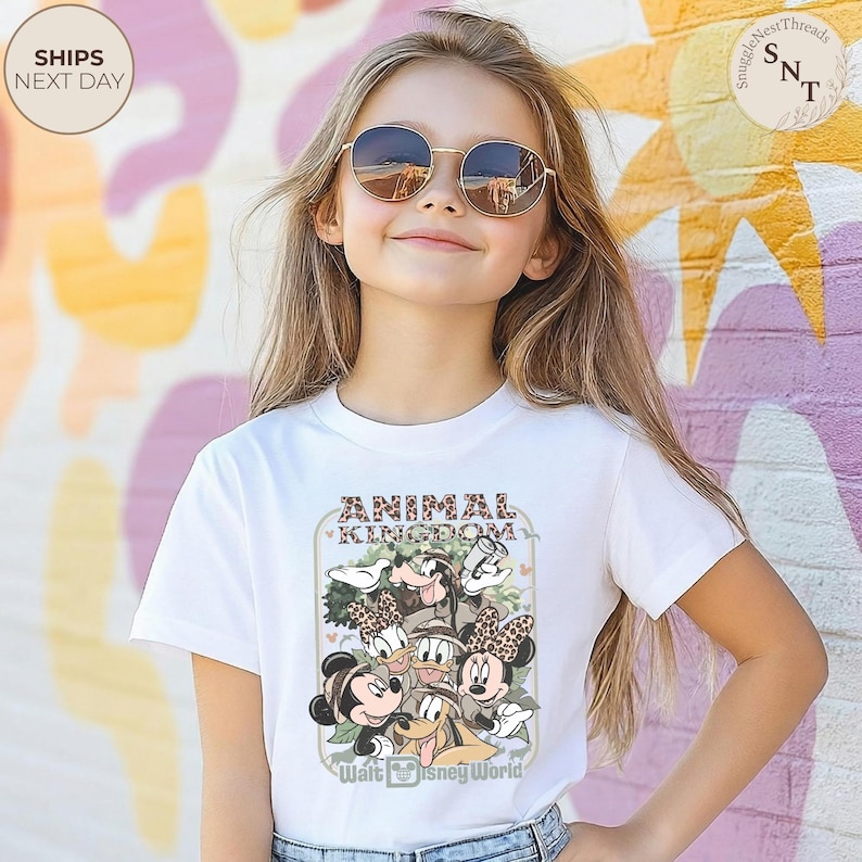 Pu&ograve; includere: T-shirt bianca con una grafica di personaggi Disney, tra cui Topolino e Minnie, Pippo e Paperino, con la scritta "ANIMAL KINGDOM" e "Walt Disney World". La maglietta &egrave; indossata da una persona giovane.
