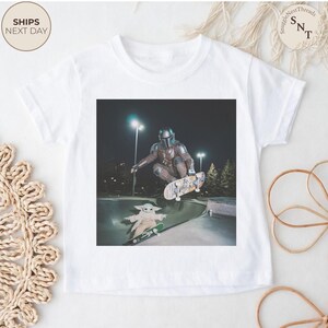 May include: White t-shirt featuring a graphic of a character in a Mandalorian helmet skateboarding, with a smaller Grogu character on a skateboard below. The image is set in a skate park at night. The shirt has the text "Ships Next Day".