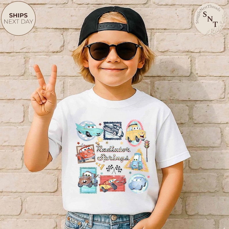 May include: White t-shirt featuring a colorful graphic of cartoon cars in stamp-like designs. The text "Radiator Springs" is printed in the center. The child is wearing a black cap and sunglasses.