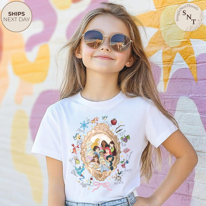 May include: A white t-shirt with a gold-framed illustration of Disney princesses. The design includes various characters, a red apple, and the word "Moana". The model is wearing round sunglasses.