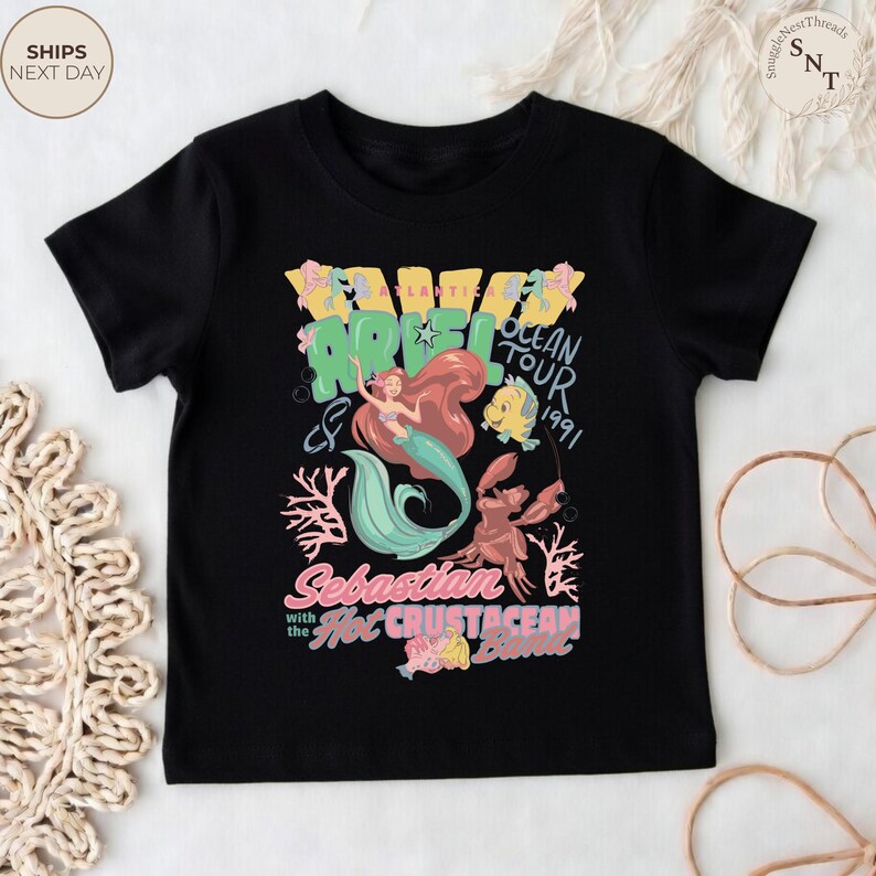 May include: A black short-sleeve t-shirt featuring a graphic print of Ariel, Flounder, and Sebastian. The text reads Ariel Ocean Tour 1991, Sebastian with the Hot Crustacean Band, and Atlantica. The design includes coral, bubbles, and musical notes.