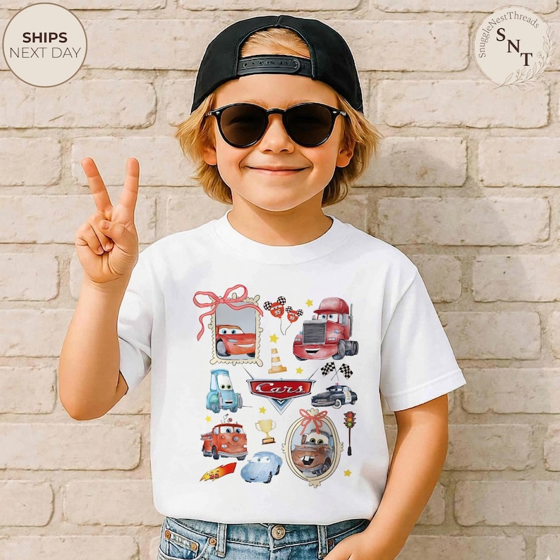 May include: White t-shirt featuring colorful watercolor illustrations of various Cars characters. The design includes images of Lightning McQueen, Mater, and other vehicles, along with the word "Cars" in a stylized font. The shirt is worn by a child.