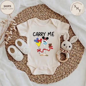 May include: Cream-colored baby onesie with the text "CARRY ME" and a cartoon character design. The design features a spoon with a face, holding balloons and a chocolate bar. The onesie is on a woven mat with baby shoes and a crochet bunny rattle.