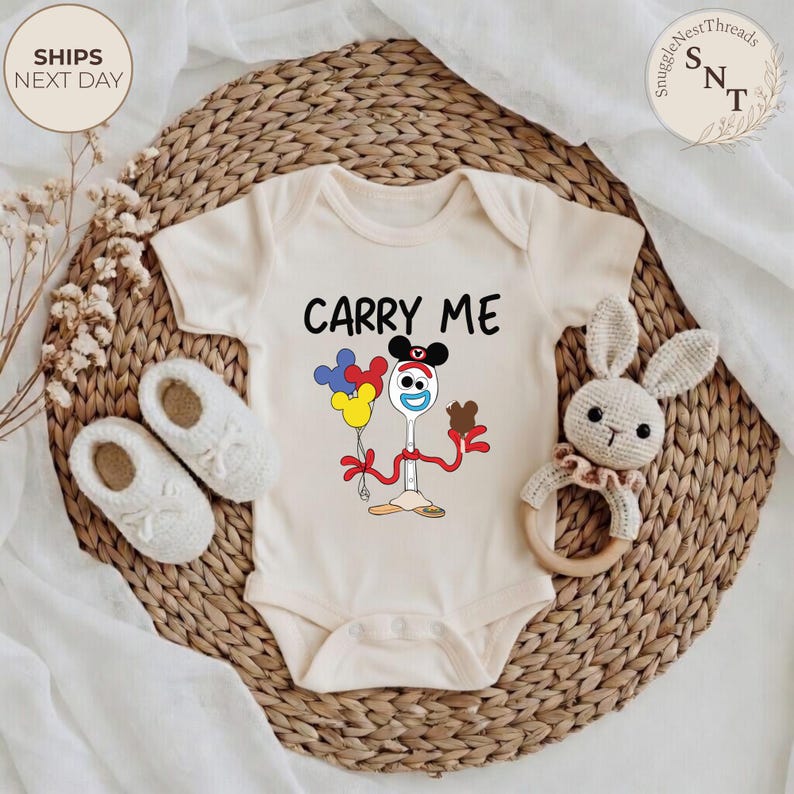 May include: Cream-colored baby onesie with the text "CARRY ME" and a cartoon character design. The design features a spoon with a face, holding balloons and a chocolate bar. The onesie is on a woven mat with baby shoes and a crochet bunny rattle.