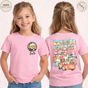May include: A pink t-shirt featuring a cartoon car design with the name "Julia" below it. The back of the shirt displays the words "Radiator Springs" with various car characters. The shirt is short-sleeved.
