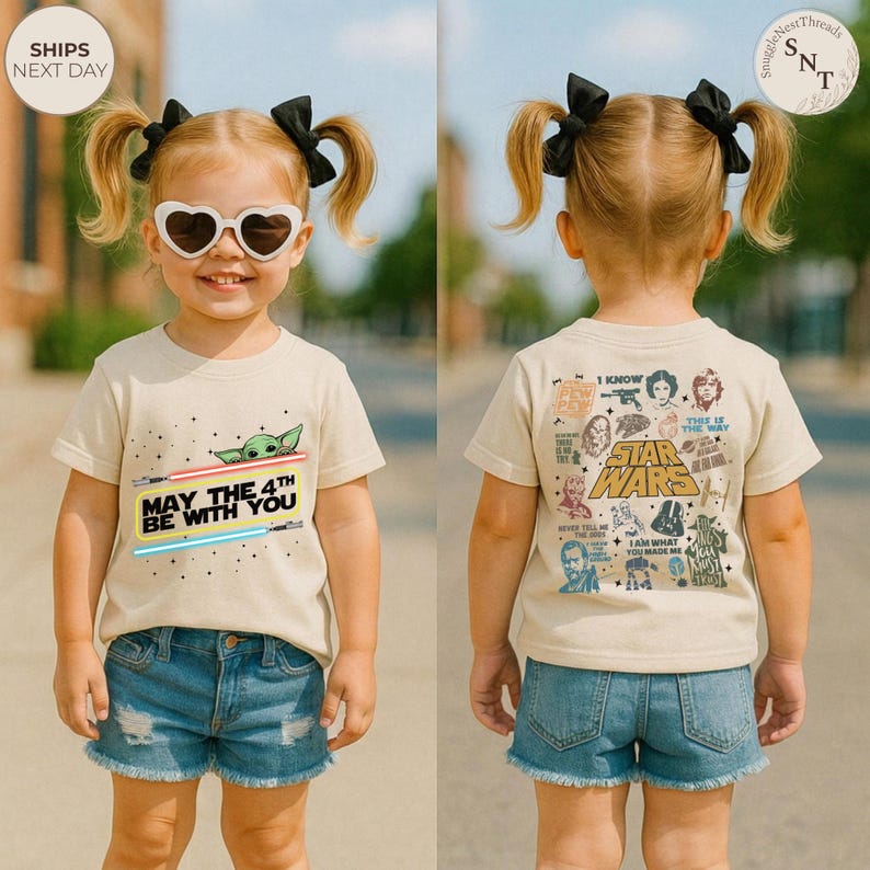 May include: A beige Star Wars-themed t-shirt with the text "MAY THE 4TH BE WITH YOU" and a graphic of Baby Yoda. The back of the shirt features Star Wars characters and the words "STAR WARS". The child is wearing denim shorts.