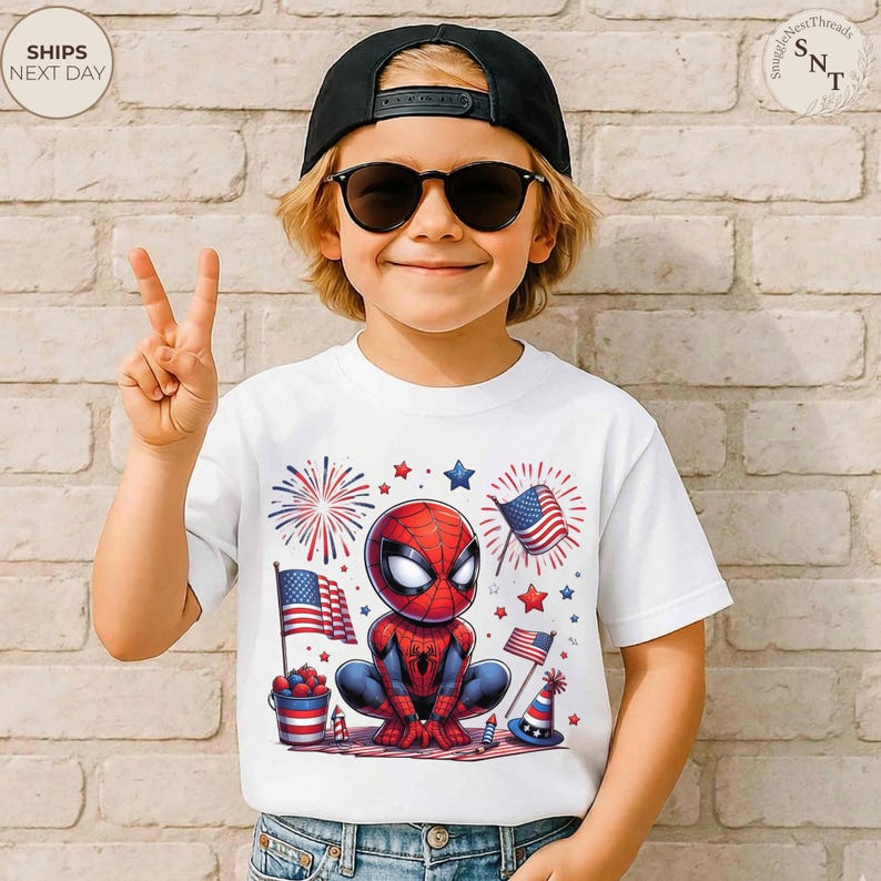 May include: A white short-sleeved t-shirt featuring a graphic of a cartoon Spider-Man character surrounded by American flags, fireworks, and stars. The design includes a bucket of berries and a festive hat, all in a red, white, and blue color scheme.