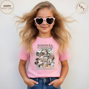 Pu&ograve; includere: Una maglietta rosa con una grafica di personaggi Disney, la scritta "ANIMAL KINGDOM" e "Walt Disney World". La maglietta &egrave; indossata da una giovane persona con occhiali da sole a forma di cuore.