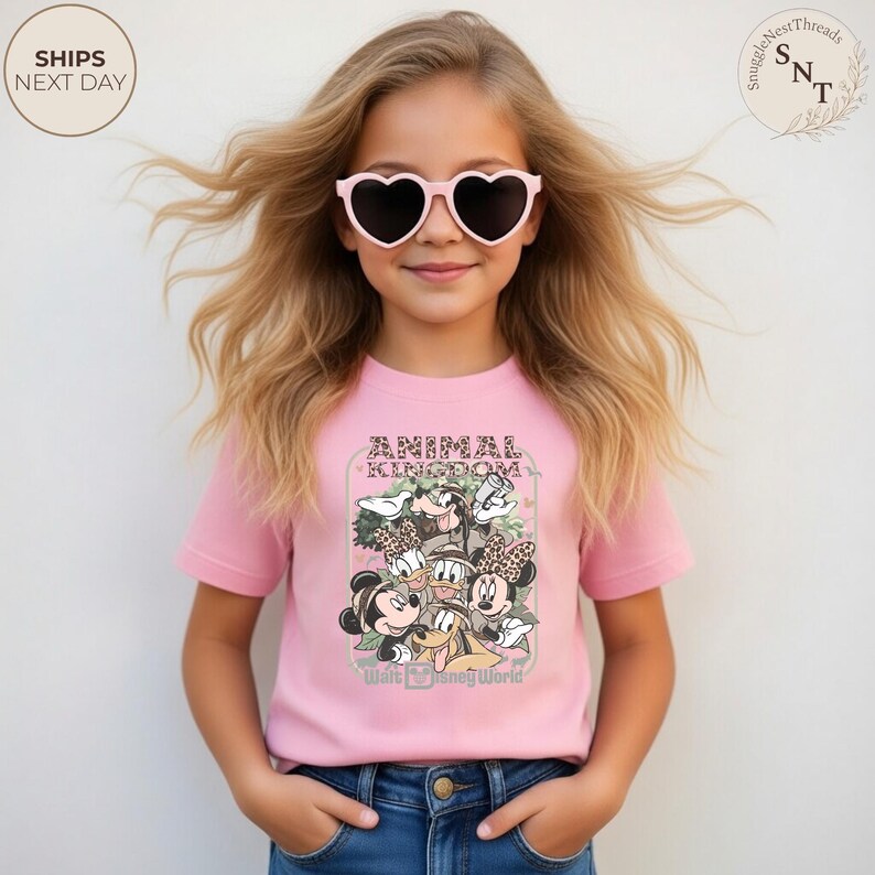 Pu&ograve; includere: Una maglietta rosa con una grafica di personaggi Disney, la scritta "ANIMAL KINGDOM" e "Walt Disney World". La maglietta &egrave; indossata da una giovane persona con occhiali da sole a forma di cuore.