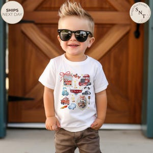 May include: A white t-shirt featuring colorful cartoon cars. The shirt has a variety of car illustrations, including a red race car, a blue truck, and a vintage car. The word "Cars" is printed in red on the shirt.