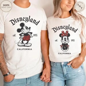 Pu&ograve; includere: T-shirt bianche sporche con grafica di Disneyland. Una maglietta presenta Topolino con pantaloncini rossi, l'altra Minnie con un fiocco rosso. Entrambe le magliette hanno stampato "Disneyland" e "California".