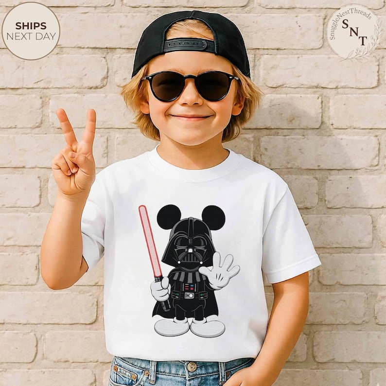 May include: White t-shirt featuring a cartoon mashup of Darth Vader and Mickey Mouse, holding a red lightsaber and waving. The shirt has a black and white design. The image also shows a child wearing a black cap and sunglasses.