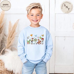 May include: A light blue sweatshirt featuring a colorful graphic of cartoon characters and balloons. The design includes a dinosaur, a cowboy, a space ranger, a horse, and a cowgirl. The sweatshirt is paired with blue jeans.