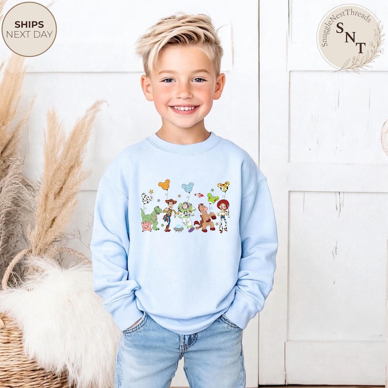 May include: A light blue sweatshirt featuring a colorful graphic of cartoon characters and balloons. The design includes a dinosaur, a cowboy, a space ranger, a horse, and a cowgirl. The sweatshirt is paired with blue jeans.