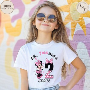 Puede incluir: Camiseta blanca con un dise&ntilde;o de Minnie Mouse, las palabras "Oh, Twoodles" y el n&uacute;mero "2" en negro con lunares rosas. El nombre "Grace" est&aacute; impreso debajo. La persona lleva gafas de sol.