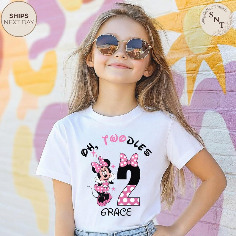 Puede incluir: Camiseta blanca con un dise&ntilde;o de Minnie Mouse, las palabras "Oh, Twoodles" y el n&uacute;mero "2" en negro con lunares rosas. El nombre "Grace" est&aacute; impreso debajo. La persona lleva gafas de sol.