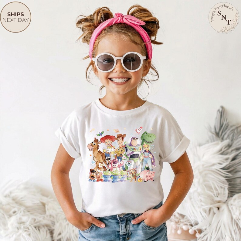 May include: A white t-shirt featuring a colorful graphic of characters from the animated movie "Toy Story". The shirt is being worn by a young person with a pink headband and sunglasses.