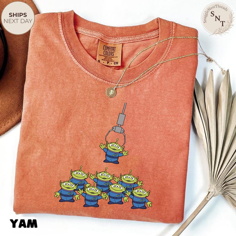 May include: A coral-colored t-shirt featuring a graphic of the Little Green Men from Toy Story. The design shows the aliens being lifted by a claw machine. The shirt has a crew neck and short sleeves. A gold necklace is draped across the shirt.