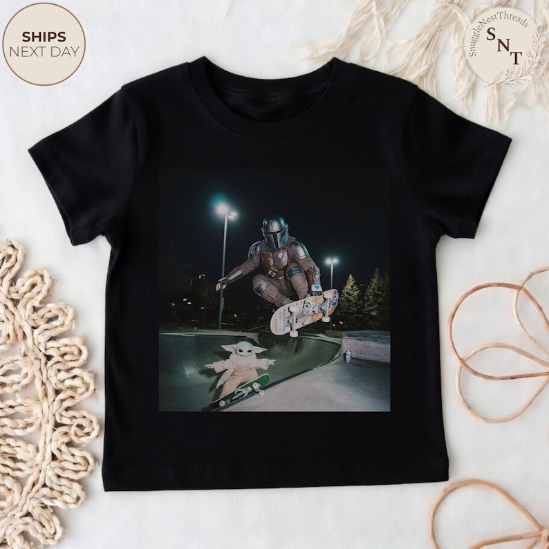 May include: Black t-shirt featuring a graphic of The Mandalorian and Grogu skateboarding in a skate park. The Mandalorian is mid-air on a skateboard, while Grogu is skateboarding below. The shirt has the text "SnuggleNestThreads" on the upper right.